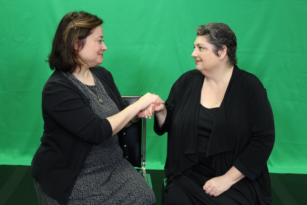 Two white women wearing formal black attire sit in front of a green screen. They are holding hands, looking into each others eyes.